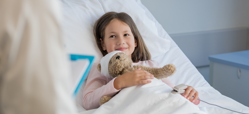 Pige ligger i en hospitalsseng med en bamse i hånden.